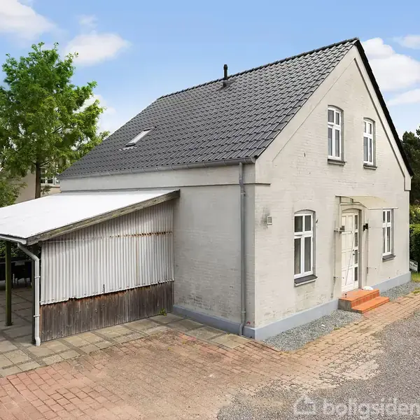 Et hvidt to-etagers hus med gråt sadeltag, placeret ved en vej. Ved siden af huset er der en carport med et bølgebliktag. Omgivet af grønt buskads og træer.