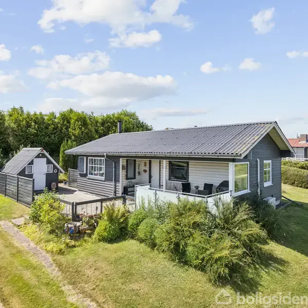 Et gråt træhus med terrasse står i en grøn have omgivet af høje buske. Der er en træsti og et skur ved siden af huset, under en klar blå himmel.
