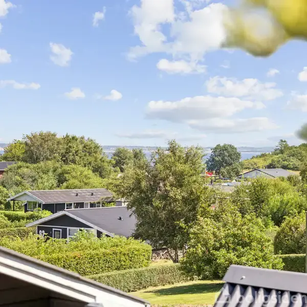 Hustag i forgrunden med flere huse omgivet af grønne træer og buske i baggrunden. Solskinsdag med blå himmel og få skyer.