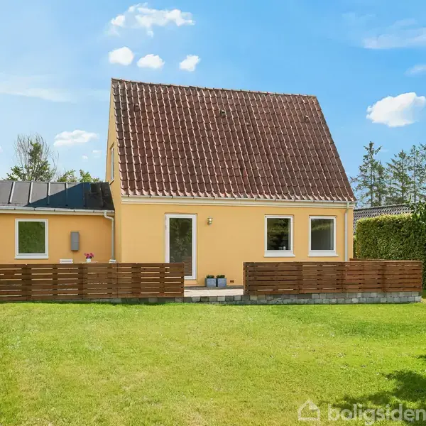 Et gult hus med rødt tag står stille i en velplejet have. Huset er omgivet af en lav træhegn, og himlen er klar med få skyer i baggrunden.