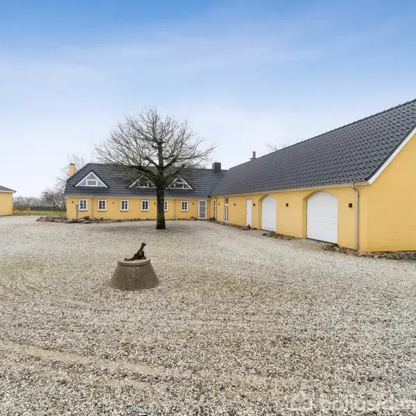 Gul bygning med mørkt tag og hvide garagedøre står i midten af en gårdsplads med en enkelt træ og en lille statue. Himmel er klar blå.