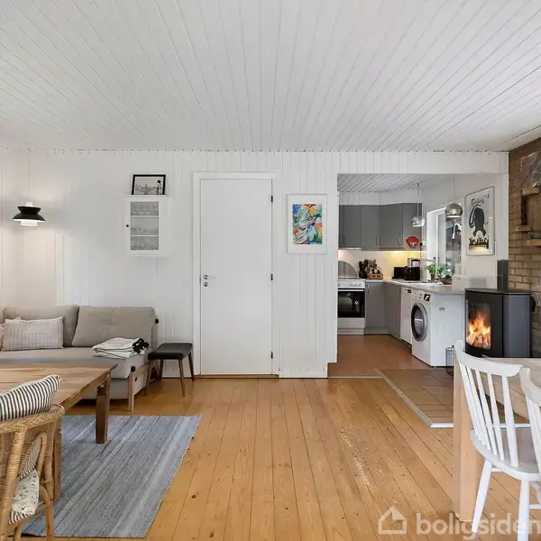 Stue med brændeovn op mod murstensvæg, træmøbler og hvide vægge. I baggrunden en åbning til køkkenet med vaskemaskine og diverse køkkenapparater. Rummets atmosfære er lys og hyggelig.