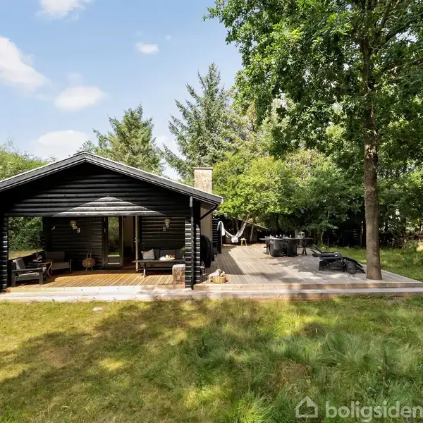 Sort træhus med overdækket terrasse står stille omgivet af græs og træer. På den åbne terrasse ses havemøbler, der giver en stemning af afslapning.