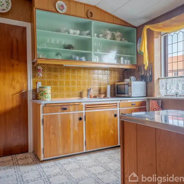 Et køkken med brune træskabe og en gul flisevæg. Hylderne har porcelæn og glas. En mikroovn og en elkedel står på bordpladen ved vinduet, som har gule gardiner.