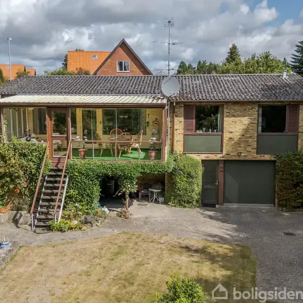 Huset består af to etager med en stor glasveranda og trappe foran. Det er omgivet af en have og en grusindkørsel, beliggende i et boligområde med andre huse.