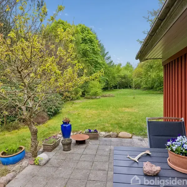 En lille terrasse med et bord og stole med blomsterkrukker på, beliggende ved siden af et rødt træhus. I baggrunden ses en stor græsplæne og grønne træer.