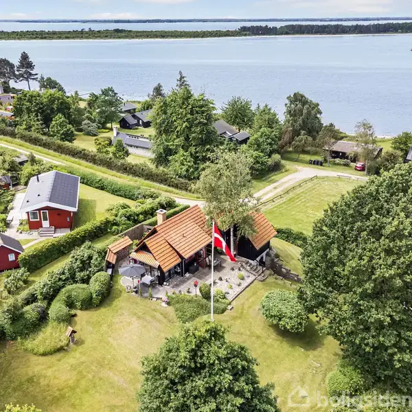 Et lille hus med rødt tag står i en grøn have med en flagstang med det danske flag. Omgivet af træer og nær en vej, med en sø i baggrunden.