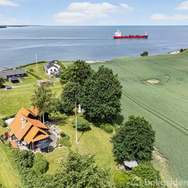 Et rødt hus med sort tag ligger i en grøn have. I baggrunden ses et stort blå hav med et rødt skib. Omgivende marker og huse fuldfører landskabet.