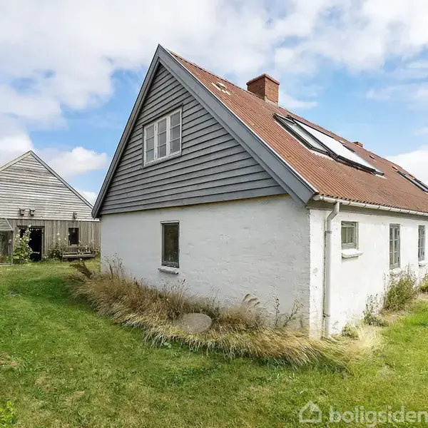 Hus med hvidkalkede mure og rødt tag med flere kviste står på en græsplæne; til venstre ses en lade og i baggrunden en silo under en blå himmel.