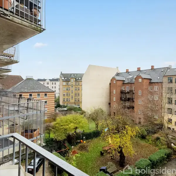 En altan med metalgelænder strækker sig ud fra en murstensbygning. I baggrunden er der flere etageejendomme og en grøn gård med træer og parkeringsplads.