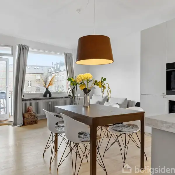 Et spisebord med blomster i vase står i et lyst køkken-alrum. Baggrunden har store vinduer med udsigt til bygninger og en balkon med grønne planter.