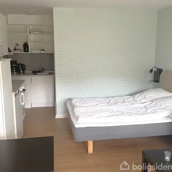 En seng står ud for en lyseblå væg i en enkelt indrettet værelse med et lille køkken i baggrunden og en blå sofa.