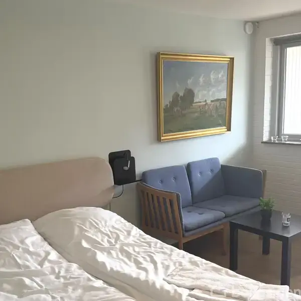 En seng står uopredt i et soveværelse ved siden af en lille sofa og et sofabord. Der hænger et maleri over sofaen, og et vindue lyser rummet op.