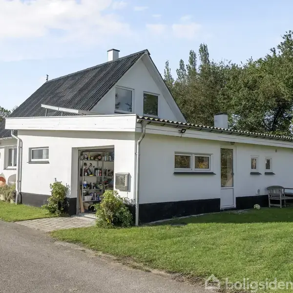 Hvidt hus med mørkt tag, omgivet af grøn have og træer. Fortil står en garage med åben port, hvor værktøj hænger. Bænke og dekorationer er placeret langs husets side.