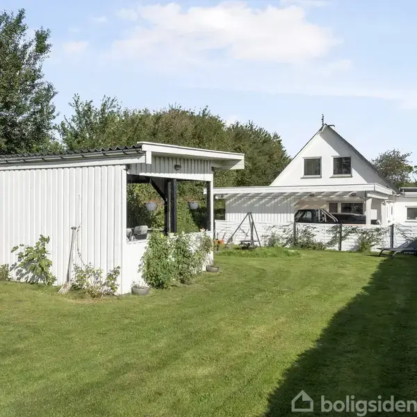 En hvidmalet garagebygning står stille i en frodig have, flankeret af en grøn hæk. I baggrunden ses et to-etagers hus med et spidst tag.