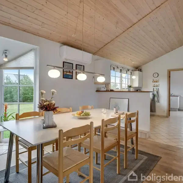 Træbord med seks stole står i et lyst køkken-alrum med skrå trælofter. Stuen har store vinduer med udsigt over en grøn have. Loftslamper hænger over bordet.