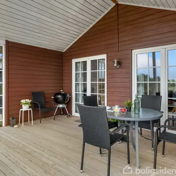 En træterrasse med sorte møbler og en grill i et hjørne. En rund bord er dækket med friske blomster og kander. Terrasse er omgivet af glasvinduer og rød facade.