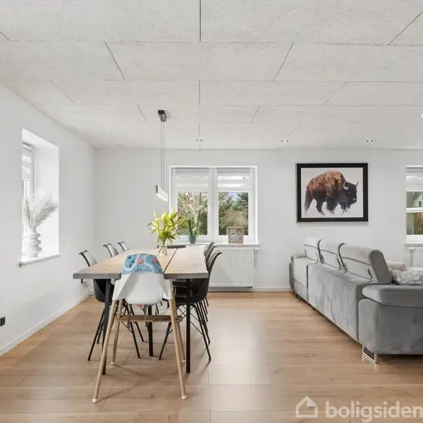 Spisebord med stole står i et lyst rum. En stor grå sofa er ved siden af med et maleri af en bison på væggen. Der er et vægur uden tal.