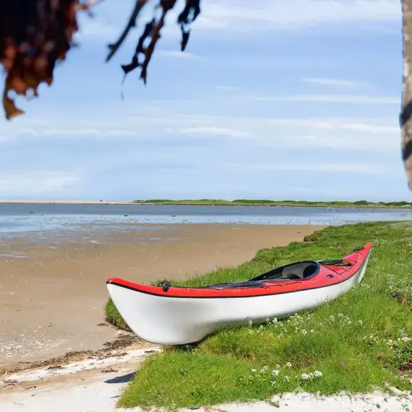 En hvid kajak med rød kant ligger stille på en grøn græsplæne nær en sandstrand ved en bred kystlinje. Himlen er lys med spredte skyer.