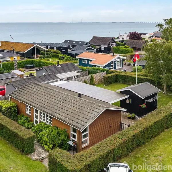 Et træhus med en asfaltbelagt tagflade omkranset af hække, ses fra oven. Huset er placeret i et kystnært sommerhusområde med udsigt over havet og flere nærliggende huse.
