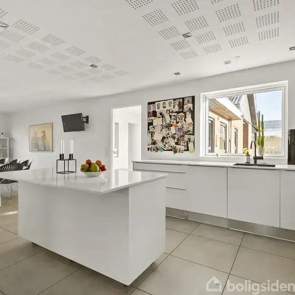 Et moderne køkken med en hvid køkkenø står centralt med frugter på toppen. Lysindfald fra vinduer, kunst på væggen, og et spisebord ses i baggrunden.