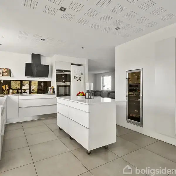 Et moderne køkken med hvidt interiør, køkkenø med frugtskål, indbygningsovne. Vægmonterede skabe og store vinduer giver naturligt lys. Flisegulv skaber en ryddelig og funktionel atmosfære.