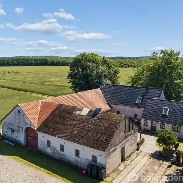 Gårdbygning med hvid murstensfacade og rødt tegltag ligger i landligt område, omgivet af grønne marker og træer under en blå himmel med få skyer.