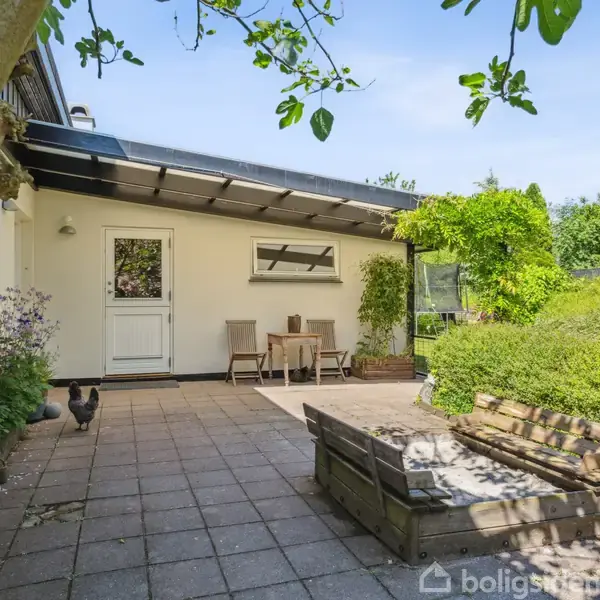 Hus står ved en terrasse med fliser, hvor to stole og et bord er placeret. En høne går rundt i haven, omgivet af grønne planter og træer under en blå himmel.