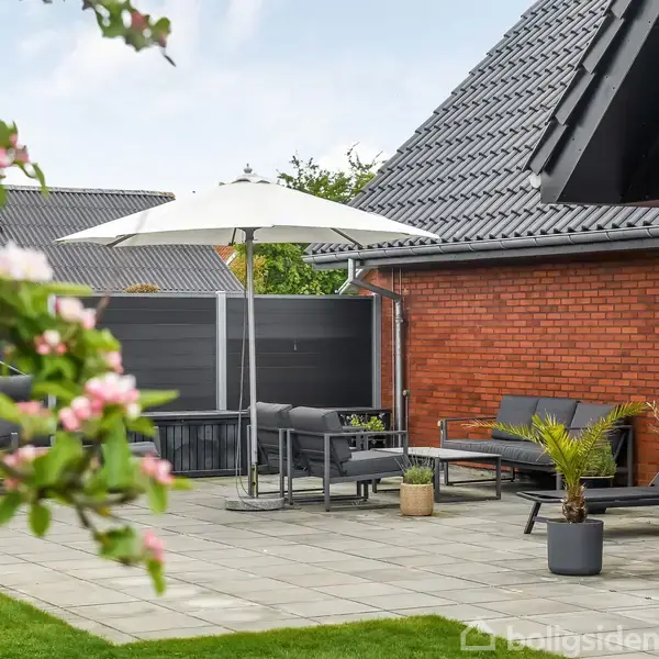 En terrasse med sorte møbler og en stor hvid parasol står ved siden af et rødt murstenshus. Omgivet af grønne planter og blomstrende grene.