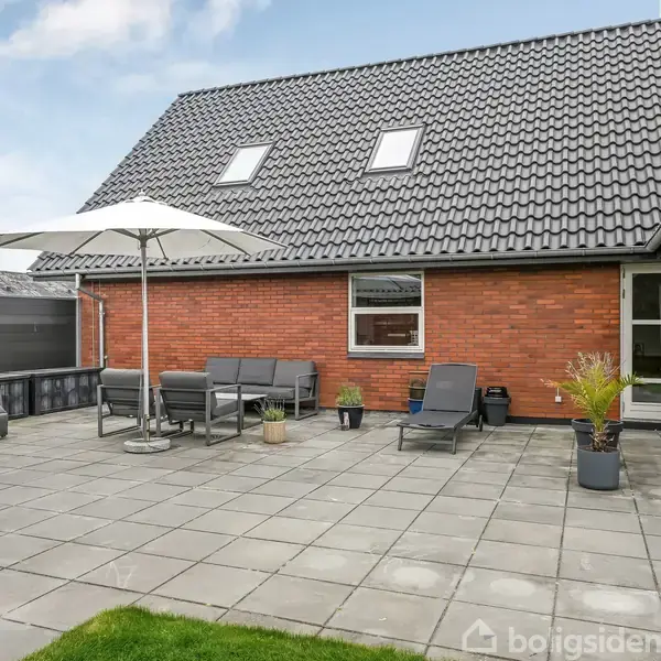 Huset har en rød murstensfacade med mørkt tag, og terrassen er møbleret med grå loungemøbler og en parasol. Omkringliggende område har fliser og grøn græsplæne.