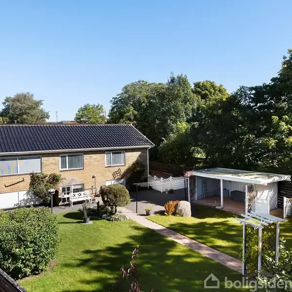Et ensidet hus med sort tag står i en velplejet have med græsplæne, træer og en flisebelagt sti. Til højre ses en hvid pergola og en lille terrasse.