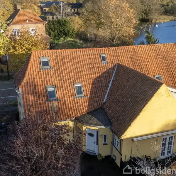 Gult hus med rødt tegltag ses ovenfra, omgivet af træer med efterårsblade. I baggrunden er der en sø og flere huse. Vej løber langs venstre side.