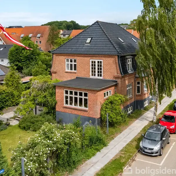 En rød murstensvilla med sort tag ligger i en grøn have, omgivet af træer og buske. En dansk flagstang står foran huset. Biler er parkeret langs vejen ved siden af.