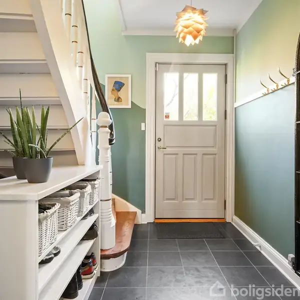 En entre med flisegulv og grønne vægge. En sko-reol med plante står under en trappe. En spejlet væg til højre, og en hvid hoveddør for enden. Over trappen hænger to billeder og en lampe.