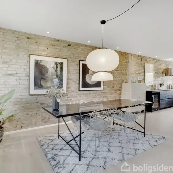 Et spisebord står i midten med to stole omkring. En stor lampe hænger over bordet. Baggrunden er en murstensvæg med kunstværker. I siderne er køkkenelementer synlige.