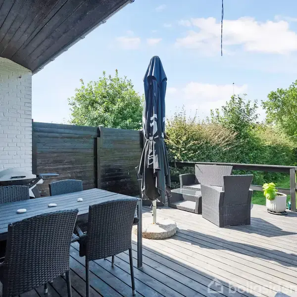 En terrasse med et spisebord og seks stole står under en sort parasol. Der er en grill ved siden af en hvid murstensvæg. Baggrunden viser en grøn have.