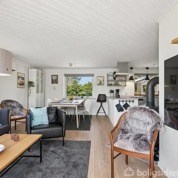 Et gult murstenshus med terrasse i en frodig have nær en kyst. Huset har store vinduer, og marker samt træer omkranser området. Havet kan ses i baggrunden.