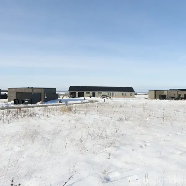 Flere moderne, lave bygninger står på en snedækket mark i et åbent landskab under en lyseblå himmel.
