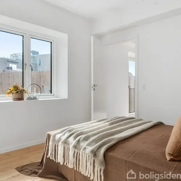 Et soveværelse med en brun seng dækket af en stribet plaid og brun pude, står i et lyst rum med hvide vægge. Ved vinduet står planter i potter. Dør åbner til anden del af huset.