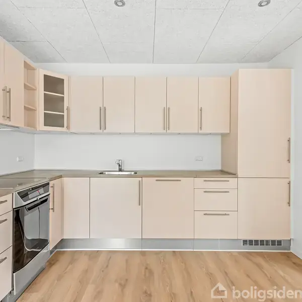 Køkken fyldt med lyse skabe og skuffer arrangeret langs væggen. Der er en sort ovn og indbygget vask. Gulvet er lyst trægulv, og væggene er hvide.