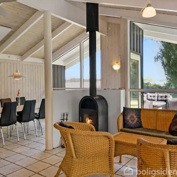 En pejs opvarmer stuen, møbleret med kurvestole og sort sofa. Til venstre står et spisebord med sorte stole, mens store vinduer åbner ud til en terrasse med udesofaer.