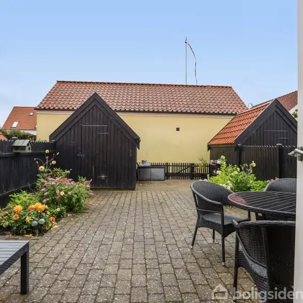 Terrasse med sorte kurvemøbler og blomsterbede på hver side, set mod lyse gule skure med røde tage. Døren til terrassen er åben, afslørende en klar himmel.