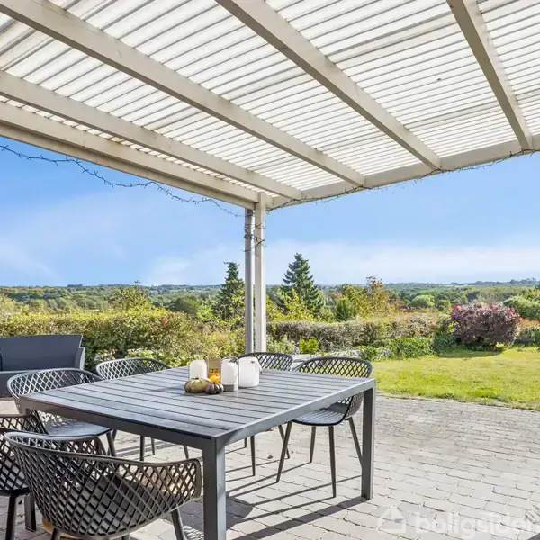Et havebord med stole står under en overdækket terrasse. Bordet har dekorationer, og terrassen har udsigt til en frodig have med grønne buske og en åben blå himmel.
