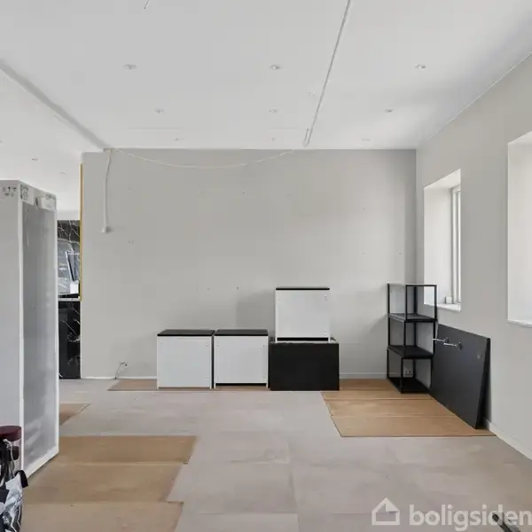 Et rum under renovering med uåbnede møbelkasser og værktøjer. Rummet har hvidmalede vægge og flere store vinduer, der giver naturligt lys ind i rummet.