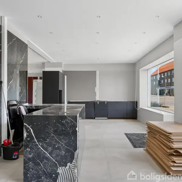 Et rum med sorte marmorlignende vægge og bardisk, en stabel træplader til højre. Store vinduer giver naturligt lys, omgivet af en moderne bygningsfacade udenfor.