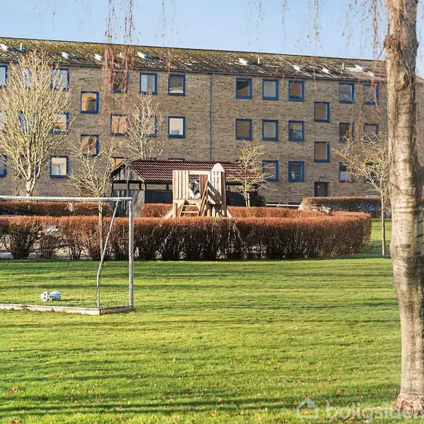 En legeplads med en klatrestativ står på en grøn græsplæne, omgivet af hække og træer, foran en stor murstensbygning med mange vinduer.