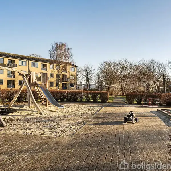 En legeplads med en stor gyngestativ og rutsjebane står på sand. En lille sort legetøjsbil er placeret på flisebelægning. Baggrunden viser et murstensbyggeri og træer.