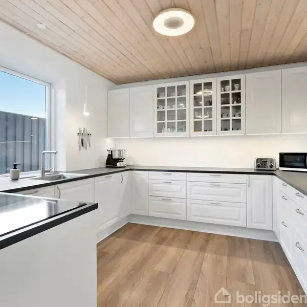 Et moderne køkken med hvide skabe og sorte bordplader. Et vindue lader naturligt lys komme ind. Rummet har træloft og trægulv, med en mikrobølgeovn og kaffemaskine på bordet.