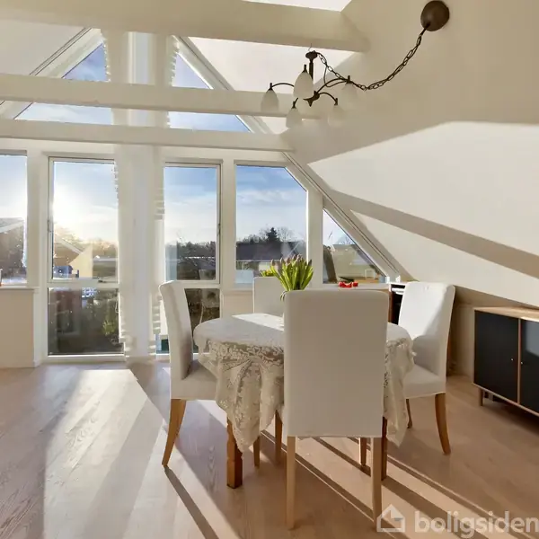 En lys spisestue med et bord og fire stole. Sollys strømmer ind gennem store, trekantede vinduer, og et skab med bøger står til højre. En lampe hænger i loftet.