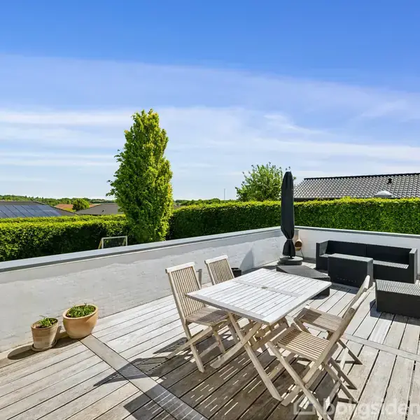 Terrasse med træbord og stole, omkranset af hvid mur. Der står en sort parasol og sofa i hjørnet. Grøn hæk og træer i baggrunden under en klar blå himmel.